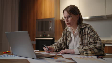 Portrait-of-a-woman-with-glasses-working-remotely-in-a-home-office-at-a-desk-with-a-laptop-and-notes-data-on-a-graph-business-economist-analyst
